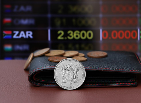 The heap coins of South Africa Rand money and black leather wallet on brown floor with digital board of currency exchange money background.