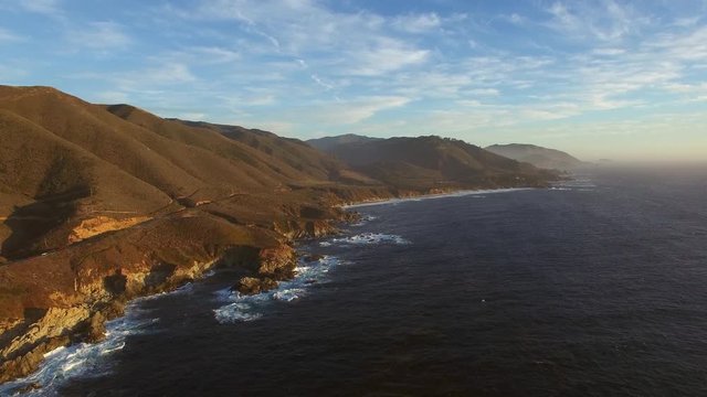 Aerial: Waves in sea by coastal road on rocky mountains against sky during sunset - Big Sur, California