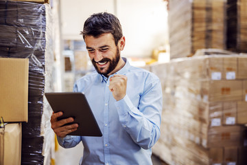 Young happy bearded supervisor standing in warehouse and cheering for good salary while holding tablet and looking at it.