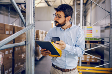 Young handsome bearded employee holding tablet and smart phone in hands and trying to finish work...