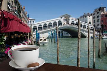 Fototapeta na wymiar Cup of back coffee or tea with cookie with view on famous Canal Grande and Rialto Bridge in Venice, Italy