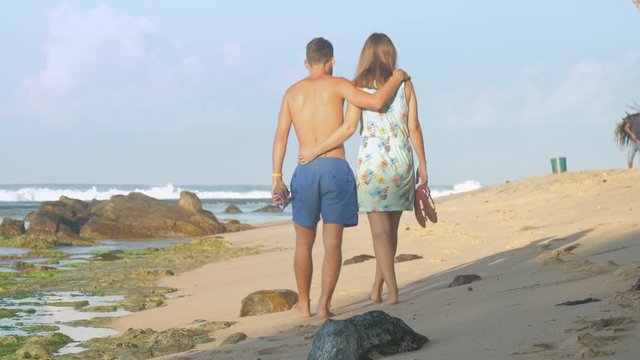 lovely couple walks barefoot along wet beach covered with green seaweed grown in rocks backside view slow motion. Concept exotic travel oceanscape environment