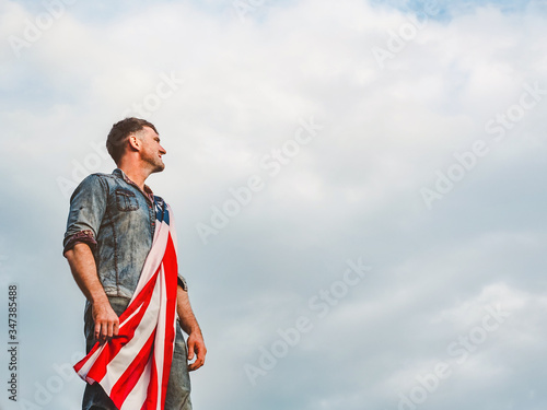 Attractive man holding Flag of the United States on blue sky background on a clear, sunny day. View from the back, close-up. National holiday concept
