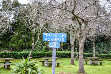 Old blue sign with text Proibido Acampar in Portuguese language. TRANSLATION: Camping forbidden. Park with green trees, benches and tables in the background. No camping sign, no overnight stay.