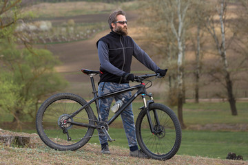 Obraz premium Sports brutal bearded guy on a modern mountain bike. Cyclist on the green hills in the spring.