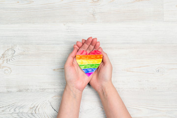 Female hands hold rainbow LGBT heart on a  light wooden table.