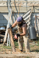 A middle-aged bearded man in a bandana cuts logs with an ax. Brutal in overalls does the hard work on a hot day.