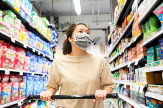 Asian Female Wearing Protective Face Mask With Shopping Cart At Supermarket Store. People Lifestyle During Coronavirus Or COVID-19 Crisis. Customer Woman Looking For Food And Grocery Stuff In Shelf