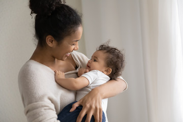 Happy loving young african American mother hold in arms sing lullabies to little toddler baby,...