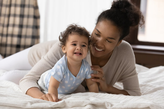Smiling biracial young mother lying relaxing on bed at home creeping with small toddler infant child, happy african American mom rest in bedroom, playing with little baby, childcare, family concept