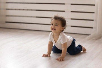 Happy small biracial baby girl child creep on all fours on warm wooden floor at home, smiling cute...