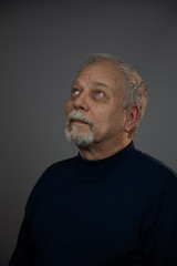 thoughtful blue eyed old man with grey beard and hair looks up standing in studio close view. Concept man emotion