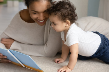 Happy young african American mom and little baby toddler relax at home bed reading children book...