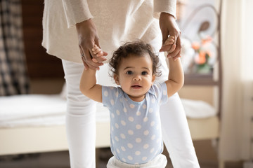 Portrait of cute small african american baby girl learn walking at home holding mom hands, little biracial toddler infant child make do first steps with mother support, childcare, upbringing concept - Powered by Adobe