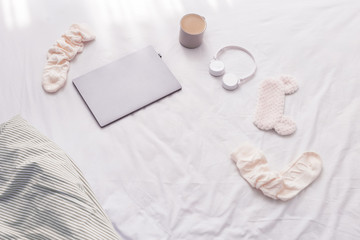 Work at home, rays of morning sun and randomly scattered things on bed. Laptop, warm socks, cup of coffee, wireless headphones, mask for sleeping on bed sheet. Top view and copy space