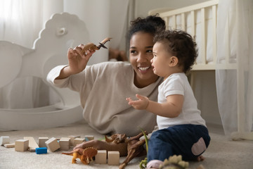 Happy biracial young mom sit relax on floor at home playing with little baby girl with toys,...
