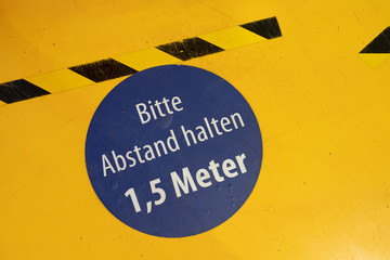 Ein Schild auf dem Boden mit Absperrband auf dem in Deutsch steht, dass man einen Abstand von 1,5...