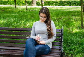 beautiful young girl in glasses writes with a pen in a notebook while sitting on a bench in a park.