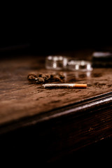 A rolled marihuana joint ready to smoke on the table with weed buds and grinder in the background. Joint is a recreational drug and a medicinal therapy as well. It is called roach, spliff, fatty...