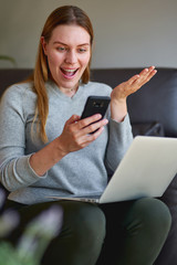 Happy woman sitting on sofa with laptop and talking on phone at home. College student studying on laptop and using phone.