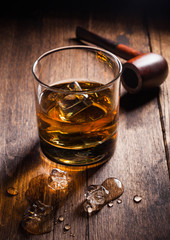 Single malt scotch whiskey in classic glass with ice cubes and smoking pipe on wooden background