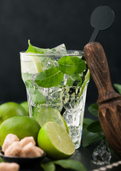 Glass of Mojito cocktail with ice cubes mint and lime on black board with spoon and fresh limes and cane sugar with wooden squeezer and steel shaker with ice.