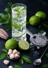 Fototapeta premium Highball glass of Mojito cocktail with ice cubes,mint and lime on black board with spoon and strainer and fresh limes with cane sugar and wooden squeezer and black shaker.
