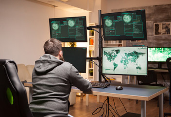 Hacker writing his hacking code on computer with multiple screens in his apartment.