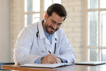 Smiling male Caucasian doctor in white uniform sit at desk fill patient form with medical history, happy man GP or physician write make notes in journal of client anamnesis, healthcare concept