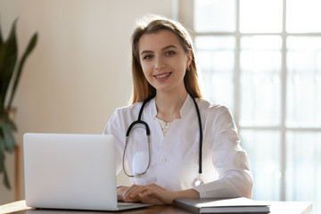 Portrait of successful young female doctor in white medical uniform and stethoscope sit at modern workplace with laptop, smiling millennial Caucasian woman nurse or GP work on computer in hospital