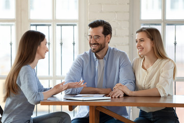 Happy young couple talk consult with female real estate agent or broker buying first house together, smiling millennial husband and wife discuss moving to new rent home with woman realtor at meeting