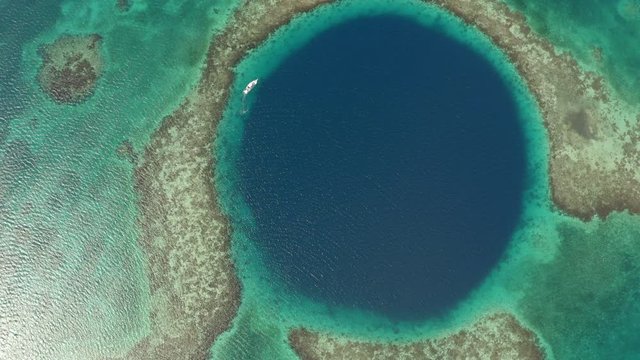 Aerial top view of yacht in famous marine sinkhole, seascape on sunny day while drone turning from right to light - Great Blue Hole, Belize
