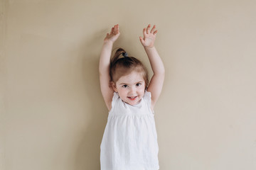 A happy little girl of European appearance stands on a beige background. Three year old baby in a white t-shirt. hands up