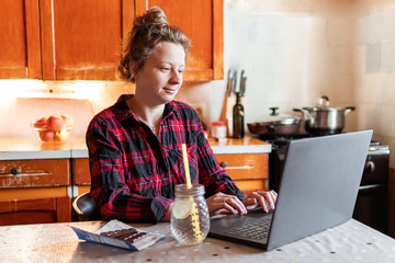 Young smiling woman is working on her laptop at home at the kitchen, drinking water with lemon and...