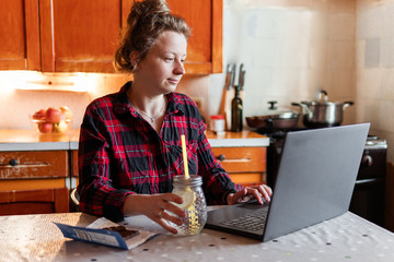 Young smiling woman is working on her laptop at home at the kitchen, drinking water with lemon and...