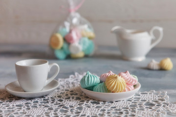 Multi-colored meringues in pastel shades with cups and a jug on a light textured wooden background. The concept of Breakfast with tea or coffee, home comfort and tenderness. Close-up, selective focus