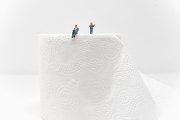 A businessman working on a laptop perches on top of a roll of toilet paper while a businesswoman presents in the background - Tiny People Working From Home