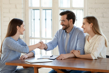 Smiling young couple shake hand greeting getting acquainted with female realtor or designer at meeting, happy millennial husband and wife handshake woman consultant closing deal in office