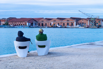 Tourists sitting on white chairs viewing the blurred industrial shipyard along the sea in Venice,...