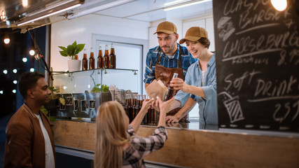 Food Truck Employee Hands Out Burger and Cold Drink to Happy Young Hipster Customer. Successful Commercial Truck Selling Street Food in Modern Cool Neighbourhood.