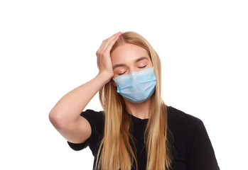 Woman wears protective white mask against virus, feels strong pain in temples, holding fingers to head. Young girl with long blonde dressed in black t-shirt