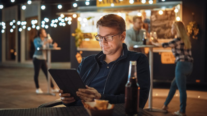 Handsome Young Man in Glasses is Using a Tablet while Sitting at a Table in a Outdoors Street Food Cafe and Eating Fries. He's Browsing the Internet or Social Media, Posting a Status Update. 