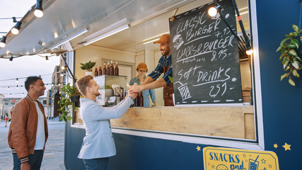 Food Truck Employee Hands Out a Freshly Made Gourmet Burger to a Happy Young Man in a Suit. Street Food Truck Selling Burgers in a Modern Hip Neighbourhood.