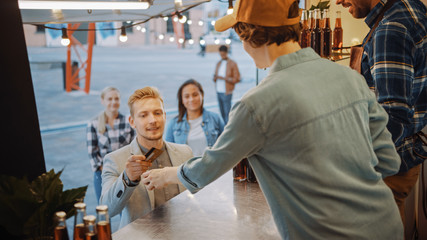 Food Truck Employee Hands Out a Freshly Made Burger to Happy Young Man in a Suit. Male is Paying for Food with Contactless Credit Card. Street Food Truck Selling Burgers in a Modern Hip Neighbourhood