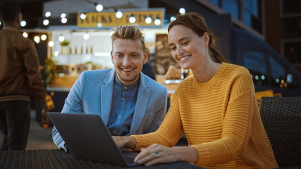 Beautiful Young Couple is Using a Laptop while Sitting at a Table in an Outdoors Street Food Cafe. They're Browsing Internet or Social Media, Posting a Status Update. They are Happy and Smile.