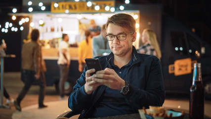 Handsome Young Man in Glasses is Using a Smartphone while Sitting at a Table in a Outdoors Street Food Cafe and Eating Fries. He's Browsing the Internet or Social Media, Posting a Status Update. 