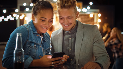 Beautiful Young Couple is Using a Smartphone while Sitting at a Table in an Outdoors Street Food Cafe. They're Browsing Internet or Social Media, Watching Videos. They are Happy and Smile.