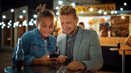 Beautiful Young Couple is Using a Smartphone while Sitting at a Table in an Outdoors Street Food Cafe. They're Browsing Internet or Social Media, Watching Videos. They are Happy and Smile.