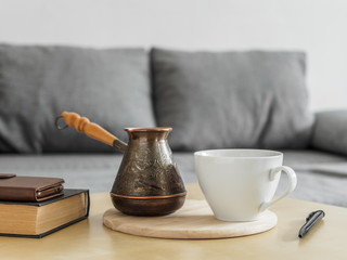 Morning coffee still life in the interior. Coffee cup, cezve and books on table, gray sofa background. Stay home concept