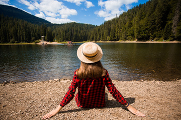 Tourist woman in hat sitting backwards and watching magic lake in mountainous landscape.